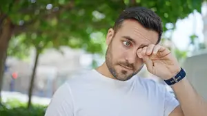 A man with a beard wearing a white shirt and a blue watch is covering his eye with his hand.
