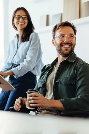 Two people sitting in a room, one is holding a cup of coffee, while the other is looking at the person holding the cup. Behind them is a wall with wooden panels, and a white wall is on the left side. The man is wearing glasses, a green jacket, and a brown shirt. The woman is wearing glasses and a blue long-sleeve shirt.