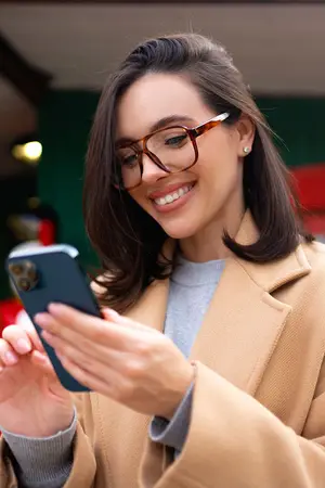 A woman wearing glasses and a beige coat is smiling and looking at her phone.