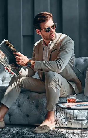 Man in a tan suit reading a magazine while sitting on a gray couch