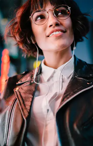 A woman wearing glasses and a leather jacket is smiling while listening to music.