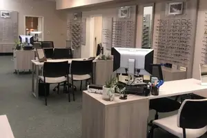 An empty room with several desks, chairs, and monitors, along with potted plants, a mirror, and picture frames on the wall.