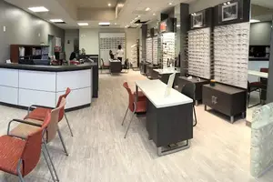 A person stands behind a counter in a retail store with chairs, tables, and eyeglass displays.