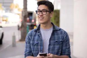 A man wearing glasses and a plaid shirt standing on the sidewalk looking at his phone