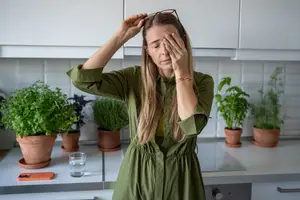 A woman in a kitchen suffering from a headache while closing her eyes