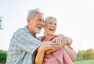 An elderly couple, an older man and woman, are standing in a field, embracing each other with happy faces under a bright sky