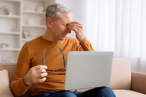 Elderly man sitting on a couch in his living room, looking at a laptop with his hand on his forehead.