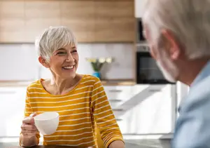 An older couple are sitting at a kitchen table and drinking coffee. The woman is smiling and holding a cup. The man is looking at her.
