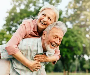An elderly couple sharing a joyful moment in an outdoor setting, with the woman playfully lifting the man as they both laugh and smile