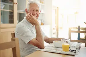 An older man is smiling while sitting at a dining table with a bowl of cereal and a glass of orange juice on the table.