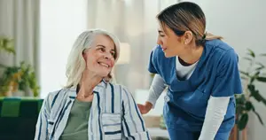 A nurse in blue scrubs is talking to an elderly woman in a striped shirt and green top in a living room with a couch, white curtains, and plants.