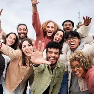 A group of people are smiling and posing for a photograph with their hands raised, while one of them is waving his hand. They are probably in an outdoor area, and some of them are wearing glasses, bracelets, and necklaces. Behind them is a blurry view of the sky with some clouds, and there is a tall structure in the distance.