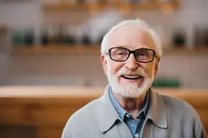 An elderly man with white hair and a beard is smiling while wearing glasses and a gray polo shirt in an indoor area, perhaps in a restaurant or bar.