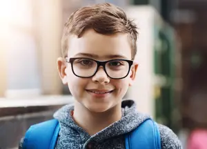 A smiling young boy wearing glasses and a blue backpack standing in front of a glass wall.