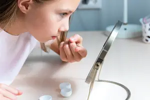 A girl trying to put a contact lens in her eye while looking in a mirror.