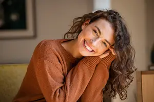 A woman with curly hair wearing a brown sweater is smiling and posing for a photo in a room with a white wall and a framed picture.