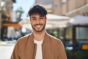 A smiling man wearing a brown jacket stands in front of a city street with blurred buildings and vehicles in the background.