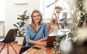 A woman sitting at a desk with a laptop, smiling and looking at the camera.