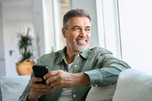 A smiling older man holding a smartphone while sitting on a couch in a room