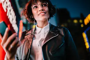 A woman with short hair wearing glasses, a black leather jacket, and a white shirt is standing outside at night while listening to music on her cell phone.
