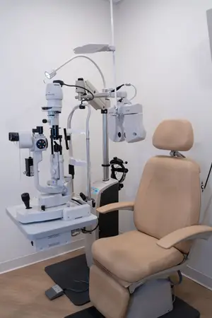 An ophthalmic examination chair is positioned in front of a wall with medical equipment nearby.