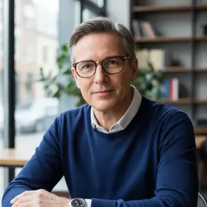 Portrait of a smiling man wearing glasses and a blue sweater, sitting in an office with a bookshelf in the background.