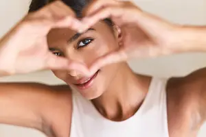 A woman making a heart shape with her hands, wearing a white tank top, and smiling while looking at the camera.