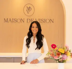 A smiling woman stands behind a desk with a flower arrangement and sign that reads 'Maison De Vision'.