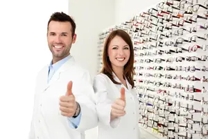 Two optometrists giving a thumbs up in front of a wall of eyeglasses