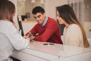 A man and a woman are looking at sunglasses at a store counter, assisted by a female employee.