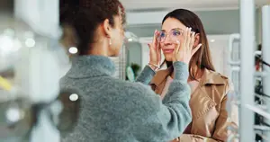 A woman wearing glasses is looking into a mirror while another woman holds her face with her hands.