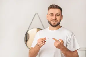A man in a white t-shirt holds a small white object in his hand, smiling, with a mirror and a wall in the background.