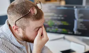 A man wearing glasses is sitting in front of a computer monitor, holding his head with his right hand and looking at the monitor, possibly feeling tired or frustrated.
