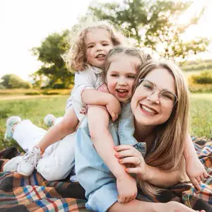 A woman and two young girls lying on a checkered blanket on a grassy field, smiling and looking at the camera