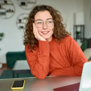 A woman is sitting at a desk, smiling, with a phone and laptop in front of her.
