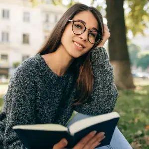 A smiling woman in glasses and a sweater reads a book in a park on a sunny day.