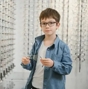 A boy is holding a pair of glasses in front of a wall with frames hanging on it