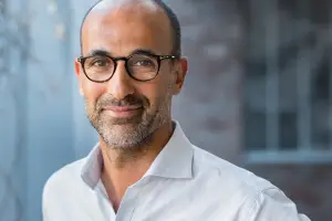 Close-up of a smiling man wearing glasses and a white shirt standing in front of a building.