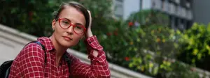A woman wearing a red checkered shirt and glasses is posing for a picture while holding her hair with her right hand.