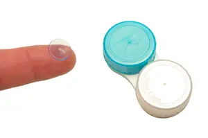 A close-up of a person's finger holding a transparent lens next to a blue and white lens case with the word Classic on it.