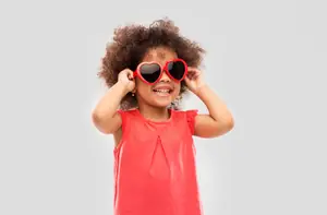 A young girl wearing heart-shaped sunglasses and a red top is smiling for a photo.