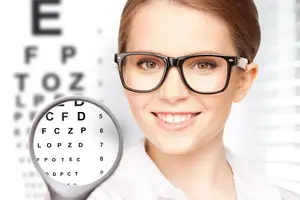 A smiling woman wearing glasses and holding a magnifying glass in front of an eye chart