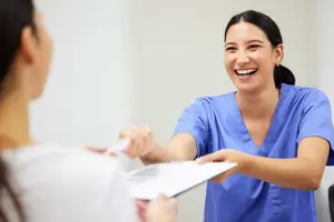 A smiling woman in a blue shirt and white smock is holding a clipboard and a pen, possibly in a hospital room, and is likely conversing with another woman who is holding a pen and a piece of paper.