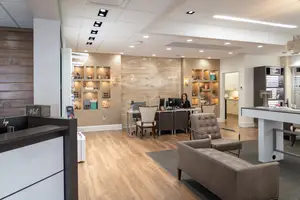 The interior of a modern office with a person sitting at a desk with a computer, surrounded by chairs, a desk, and shelves on the wall, and a gray carpet on the floor.