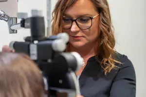 A woman in glasses is examining a young girl's eyes with an ophthalmoscope.