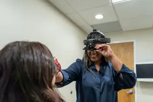 A woman is standing in a room and holding an eye testing machine on the head of another woman sitting in front of her.