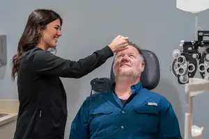 A woman is adjusting the glasses of a man sitting in a chair with an ophthalmoscope in the background.