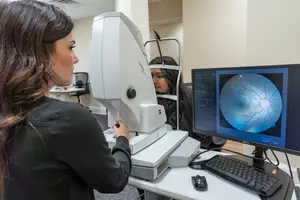 A woman wearing a black top is looking at a monitor displaying an eye scan while another woman sits behind her looking into an eye scanning device.