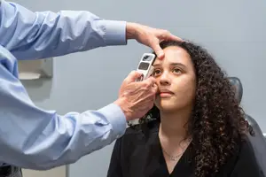 A person is sitting on a chair and is probably undergoing an eye examination. The person is wearing a black top and has curly hair. The person holding the woman's head has a blue long-sleeve shirt on. Behind them is a wall with a white toilet paper holder. The woman is looking at something in front of her.
