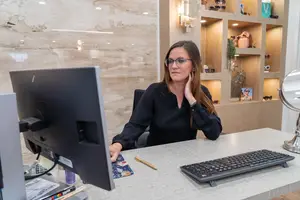 A woman is sitting on a chair and looking at the monitor while holding her phone in her left hand. She is wearing eyeglasses and a black long-sleeve shirt. A keyboard and a pen are on the desk. A mirror and a shelf with some objects are on the right. A lamp is mounted on the wall. There is a window on the left.